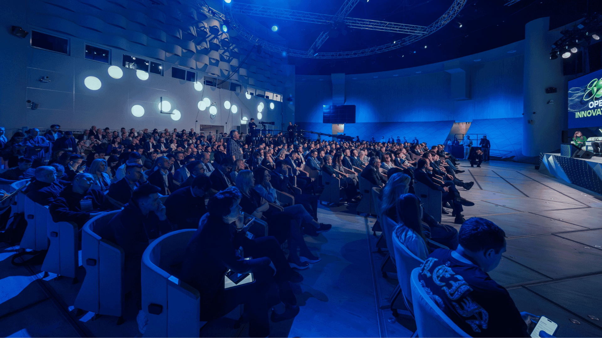 Large audience seated in a conference hall during a professional event with blue stage lighting.